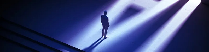 A lone person standing in dramatic light and shadow with an AI-shaped reflection projected on the ground